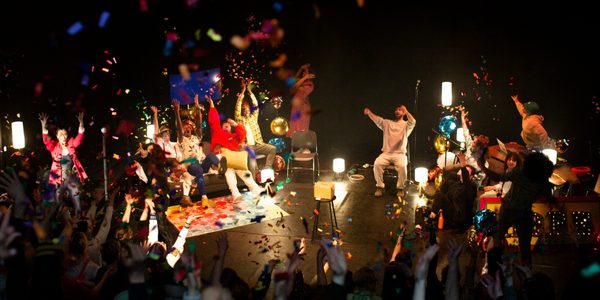 A black box theatre space with lots of people sat on stage throwing brightly coloured glitter into the air.