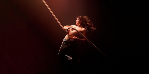 Laura Murphy is swinging on a rope that is coiled around their bare torso. They are wearing blue denim jeans. The background is dark and their is a soft light shining on them from above. Image credit Holly Revell