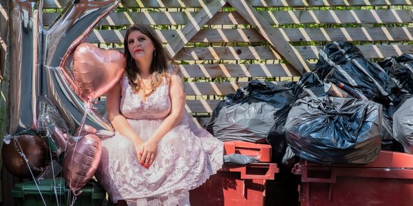 Katy Baird sitting on dumpster bins. On their left is a pile of full rubbish bags, and on their right is a collectioin of pale pink heart shaped balloons. Katy is wearing a white lace dress and white trainers.
