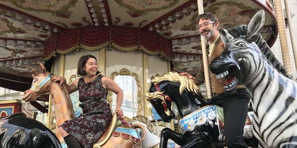 Jo and George sit on a brightly coloured, old fashioned fairground carousel. Jo is dressed in a dark red patterned dress and knee socks, sitting on a camel, smiling at George. George, dressed in dark trousers and a light yellow shirt under a brown cardigan, sits on a black horse smiling at the camera. An excited zebra is in front of him. Image by Catriona James