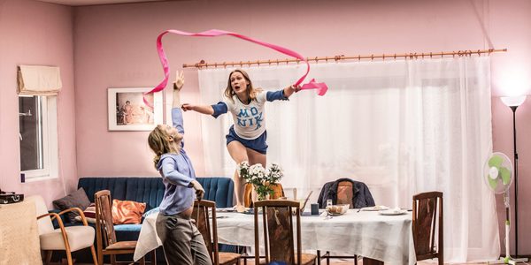 Two women in an apartment with pink wallpaper. Both are grabbing for a pink ribbon. One woman is standing on a dining room table. The other woman is reaching up with one arm to try and grab the ribbon.
