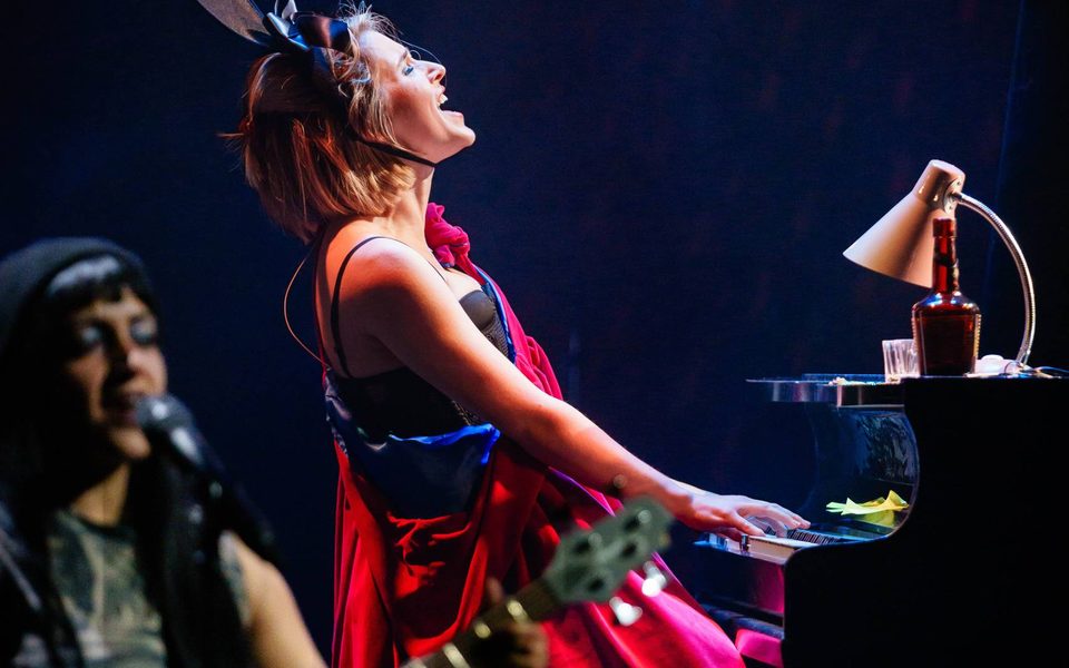 A woman in a red dress and bunny ears leans back at the piano. Light catches her face as she sings.