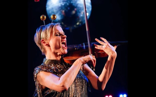 A woman with short blonde hair plays the violin passionately. Above her, a silver disco ball shimmers in the darkness.
