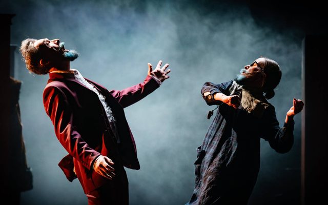 Two performers face each other and gesticulate theatrically, as if in dance. Light catches their faces. Behind them is a brooding mist.