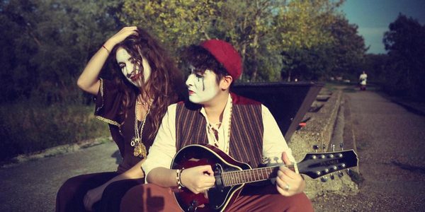 Sh!t Theatre stand in marshland, sun is glinting off their faces. Louise has short hair and carries a mandolin. Becca has long hair and is squinting. In the background you see the ghostly figure of a priest walking towards them