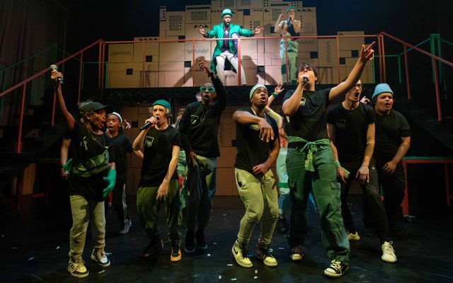A group of young people wearing black tee shirts perform in front of a cardboard structure, some of them speaking into microphones
