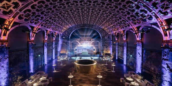 Battersea Arts Centre's Grand Hall set up for a party. White tables and chairs are positioned throughout the Grand Hall, and the ceiling is lit up in purple and blue.