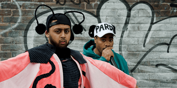 Sabir, a South Asian young man, is looking very seriously at the camera and, is dressed as the ‘Evil Butterfly’, wearing a black cap with antenna and a pink cape with black details to look like the markings on a butterfly. Nathaniel, a young black man, is behind him, wearing a teal tracksuit top and a white baseball cap with black panda ears, which reads ‘pardy’. They are both stood in front of a brick wall, covered in graffiti.