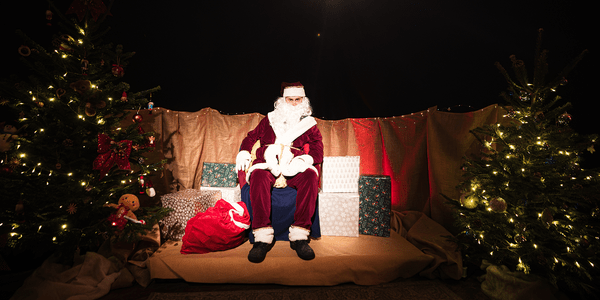 An image of Santa sat in a dark room lit by fairy light. To either side of him are decorated Christmas trees and presents.