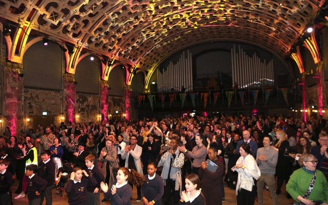 A large crowd gathers under a lattice ceiling, cast in yellow light.