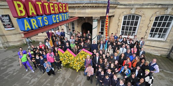 A crowd gathers outside a town hall building. They pose around a large W artwork made out of yellow flowers. A red, blue and yellow sign hangs above them, which reads: 'Battersea Arts Centre'.