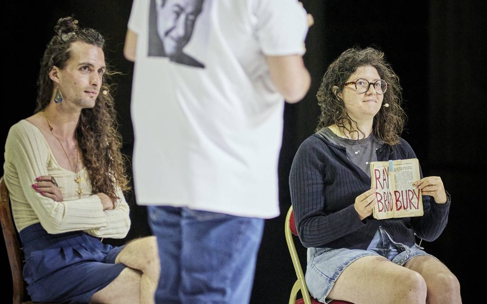 A man in a white T-shirt is stood looking over his shoulder in front of two people sat on chairs.