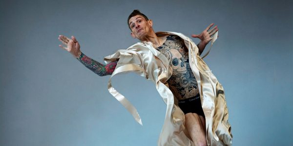 EXXY © Hugo Glendinning MH Male presenting figure, the artist Dan Daw, in mid-action pose in mid-air with arms outstretched. There is a mattress covered with a loose sheet beneath them. They are wearing a silk dressing gown that is open, exposing his tattooed skin