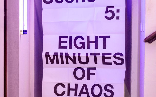Handle With Care (Ontroerend Goed) © Ans Brys A white sign with black writing on it is held up by someone, their body complete covered by the size of the sign. It reads 'Scene 5: eight minutes of chaos'