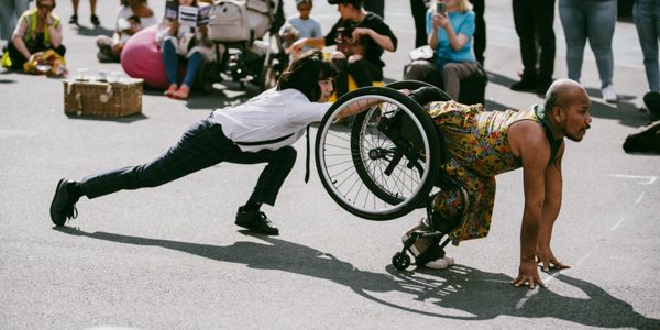 Liberty Festival © Paul Fuller Two male presenting figures who are performing outdoors. One is a wheelchair user and is learning far forward and posing on all fours while the wheelchair is still strapped to their waist. The other is behind the wheelchair user and holding onto the wheelchair with one outstretched hand, while crouching down with one long stretched behind them. There are a group of people of mixed age and gender in the background watching the performers.