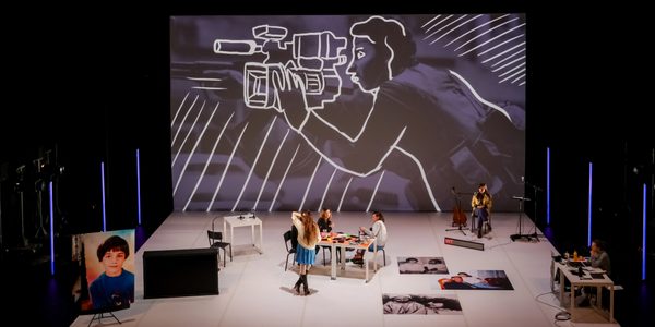 Radio Live A New Generation © Matthieu Camille A large theatre studio space with a background photo image of a person with a gun, with an outline drawing of a person with a camera superimposed on the photo. The stage is populated by 5 performers of mixed gender. There are tables, chairs, microphones, a guitar, and large photo portraits of people spread around the stage.