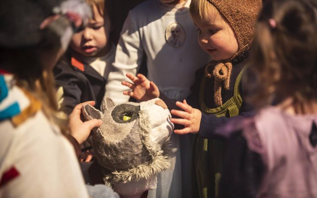 Some highly adorable toddlers are crowded round a puppet of the Yule Cat – a grey fluffy cat with a small smile at its mouth and yellow eyes. The children are all reaching out to stroke the cat.