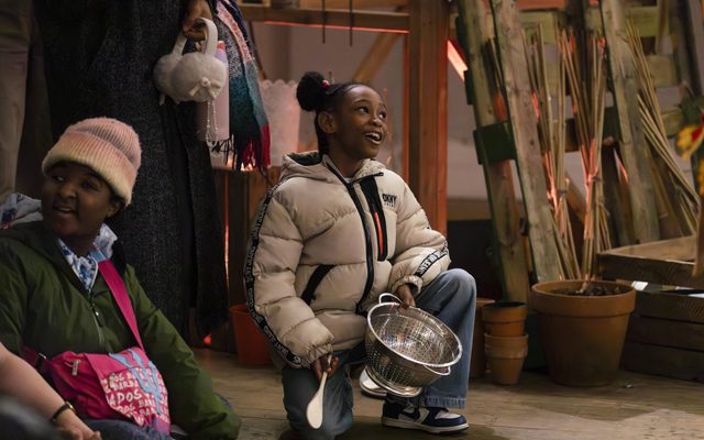 Some children are sat and kneeling on the floor smiling. One child is holding a colander in one hand and a spoon in the other, smiling off to the side. Behind her are some plant pots and some wooden crates. An adult is stood next to the child holding some white earmuffs, their face out of shot.