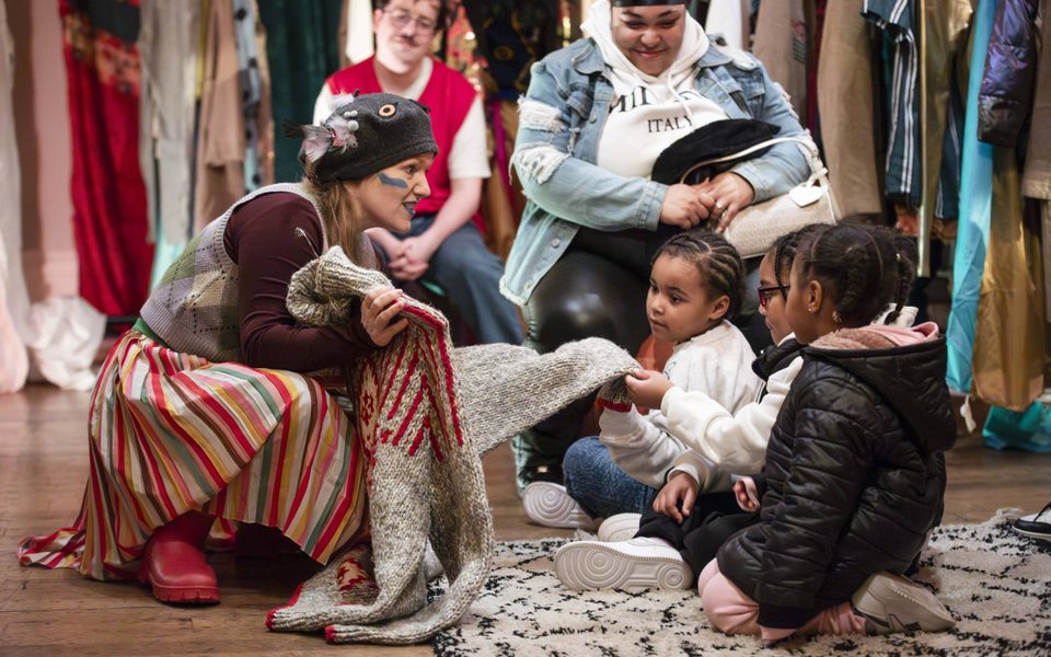 Pigeon is crouched down holding a big woolly jumper out for some small children sat on a rug to touch and look at. The children are crowded round touching the jumper. Two adults are sat behind them, looking on smiling. In the background are lots of clothes racks with different colourful garments on them.