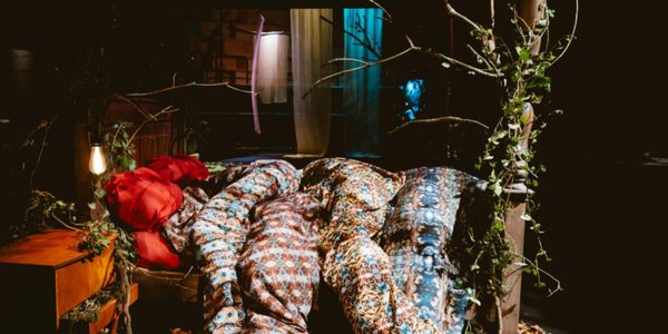 Capturing The Forest. Photo by Rosie Powell. A bedside table and wooden four-poster bed trailing with ivy, spotlit in a darkened room. Fabrics printed with ornate patterns tumble from mattress to floor, snaking like tree roots into loose autumn leaves on the surrounding floor
