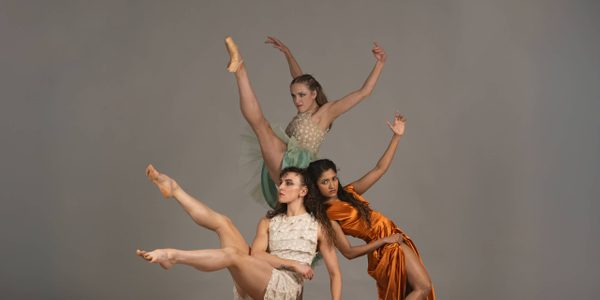 Daughters of the Sea. Photo by Krystal S Lowe. Three women, two are white and one has brown skin, surround a pile of sand against a grey background in a variety of dynamic balletic positions.