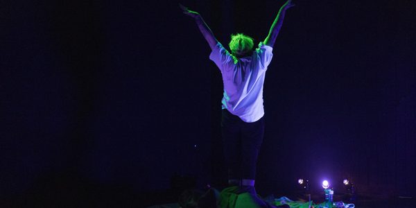 Jack, a white non-binary person with bright green hair and wearing a white t-shirt stands with their back to the camera under UV light, doing a morning stretch. They are surrounded by trinkets; a tiny pink flamingo, a book, leaves, a water bottle, a shiny pink love-heart shaped disco purse, and other nondescript things.