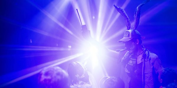 A white light is shining in the centre of the image with rays beaming off of it. Next to it is a person with large rabbit-like ears coming from their head, holding up a walkie-talkie as children look on. The whole image is washed in indigo.
