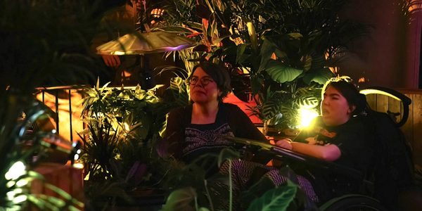 When The World Turns. Photo by Roswitha Chesher. A young woman using a wheelchair and a middle-aged woman sit beside each other. They are surrounded by wooden crates and plants, with a billowing tent canopy overhead. Atmospheric low lighting, illuminates their faces with a golden glow and casts shadows of the plant leaves around them.
