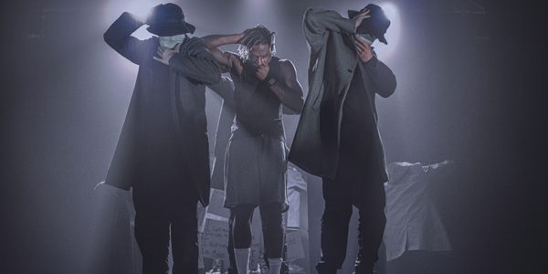 Simeon Campbell, a black man with dyed dreadlocks, wearing a black vest, grey shorts and white socks, stands centre stage, flanked by 2 other figures wearing trenchcoats, hats and masks. All 3 hold their hands over their mouths, their faces downcast. They are onstage, illuminated by spotlights.