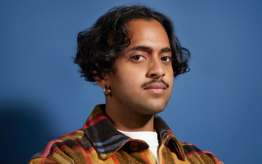 KrishnaIstha-by-Christa-Holka (2) Krishna is a brown-skinned masculine-presenting person wearing a brown and mustard plaid shirt with their arms folded, looking side on at the camera.