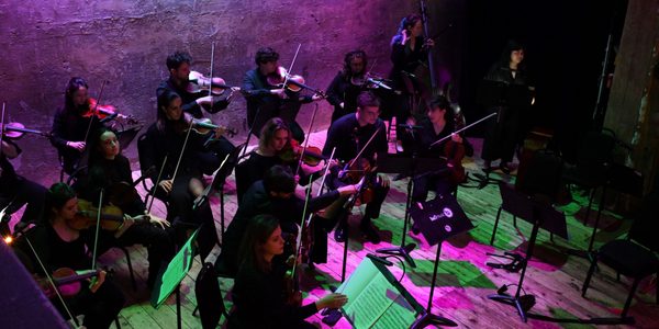 Chromatica Orchestra rehearses on stage. The orchestra is bathed in green and purple light.