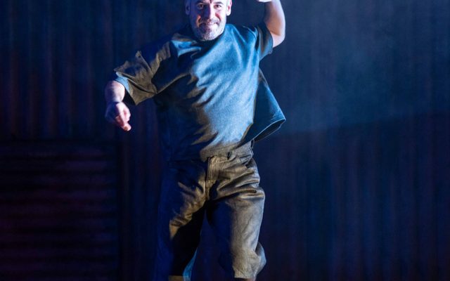 Joe Brown is dressed in grey trousers, a t-shirt and black boots. He faces the camera in the height of a jumping position; a blue light shines down from the left, illuminating his face and clothes. His arms are suspended in air and one foot off the ground. The backdrop of the stage is corrugated iron, covered by a blue haze.
