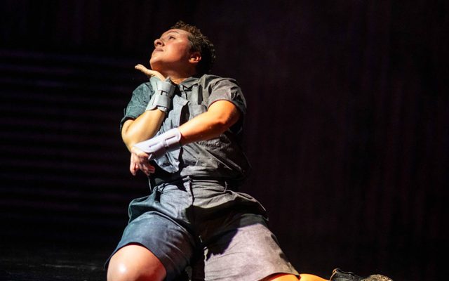 Tiiu Mortley kneels with one leg stretched out to the left on a shiny black floor. She is dressed in grey shorts and a shirt, gazing up to a soft orange light.