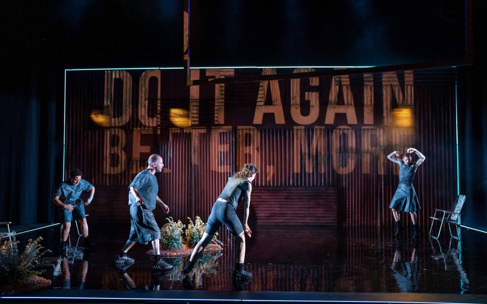A large stage with a corrugated iron backdrop that has the words 'Do it again, better, more' written across it. The cast of EXXY are on the stage, all mid-movement as they throw their arms and stomp their feet.