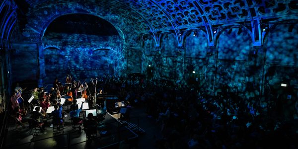 Peter and the Wolf Family Concert_credit Monika Jukobowska_MH Chromatica Orchestra on stage in the Grand Hall at Battersea Arts Centre during their The Carnival of the Animals Family concert. The hall is bathed in green and blue light which gives a ripple effect like the sea.
