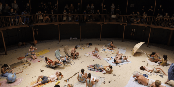 A stage with sand covering it, with people all around on twoels and in swimming costumes as if they were at the beach. The stage is in the round, and the audience is sat on a raised walkway running around the edge of the circle, peering over a balcony onto the beach scene below.