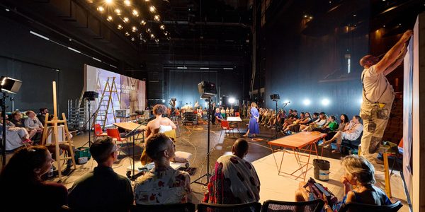 Theatre studio space with audience seated around the edge of the space. There are performers across the stage standing in various positions. There tables, chairs, theatre lamps and a projection screen spread across the space.