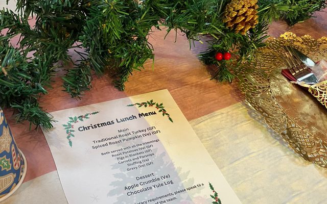 The Christmas lunch menu, next to one of the table trimmings of pine branches and holly. The menu includes spiced roast pumpkin, roast turkey, vegan apple crumble and a yule log.