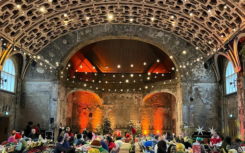 BAC’s Grand Hall with fairy lights hanging from the ceiling and long trestle tables packed with people ready to enjoy their Christmas dinner. In the back of the hall are some Christmas trees and large star decorations, with warm light projected on the walls to add the cosy atmosphere.