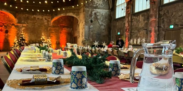 A place setting at BAC’s community Christmas meal, complete with Christmas cracker, a paper cup with a festive pattern and a jug of water. There are fairy lights twinkling from the Grand Hall ceiling and Christmas trees sparkling in the background.