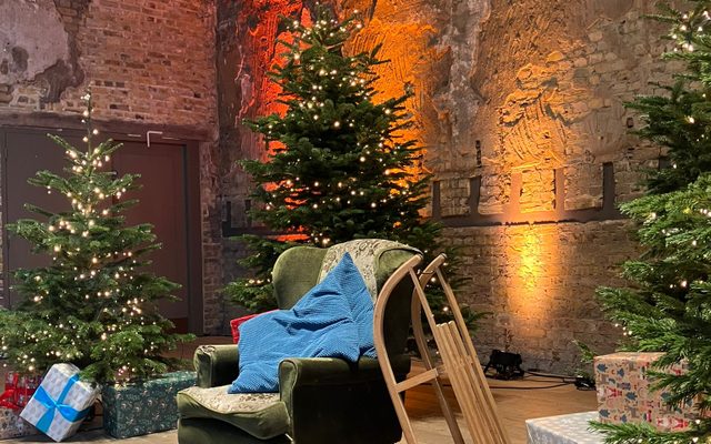 A cosy armchair with blue cushions and a sled propped up next to it, surrounded by three twinkling Christmas trees in BAC’s Grand Hall.