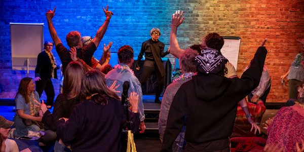 A brick wall lit by different coloured lights creates a rainbow mosaic backdrop for artist Stacy Makishi’s dance moves; a woman with a short grey bob and glasses, who is wearing a black long-sleeved shirt and black trousers. The chaotic crowd of people are joining in and display their unique poses, some with hands gesticulating, others joyfully still, sitting on the floor.