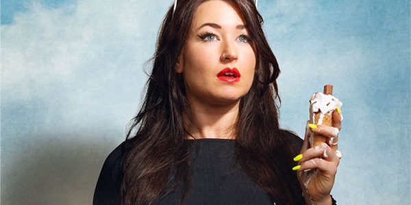A white woman with black hair wearing red lipstick is standing in front of a cloud-filled sky background. She is holding a half-melted ice-cream in her left hand and gazing upward in awe of something out of frame.