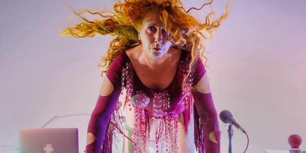 A white woman with long wavy ginger hair is kneeling on a desk with both hands down in front of her. She is wearing a dark pink long-sleeved leotard with strings of large light pink jewels and cloth tassels under the arms. There a microphone on her right and a laptop on the left.