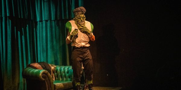 A person dressed in Frankenstein-esq prosthetics and chunky boots is standing on stage drinking a Monster energy drink. Behind them a green leather chair and teal curtains.