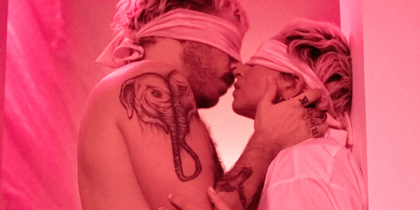 A softly lit pink room frames two people embracing for a kiss. The pair are both wearing white silk blindfolds, have various tattoos, and matching blonde hair with dark brown roots that have grown out.