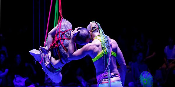 A tentative audience sitting on the floor blur into the shadows behind two vibrant bold figures, illuminated in purple light. One person is suspended in air by rope bondage knot, with neon red, pink and green rope, hands crossed behind their back. The other, with their back to the camera is gently holding the first person's head and pressing their own to it, creating a warm sense of connection and safety. In the purple background various mundane objects are spun in neon macramé creating unique patterns.