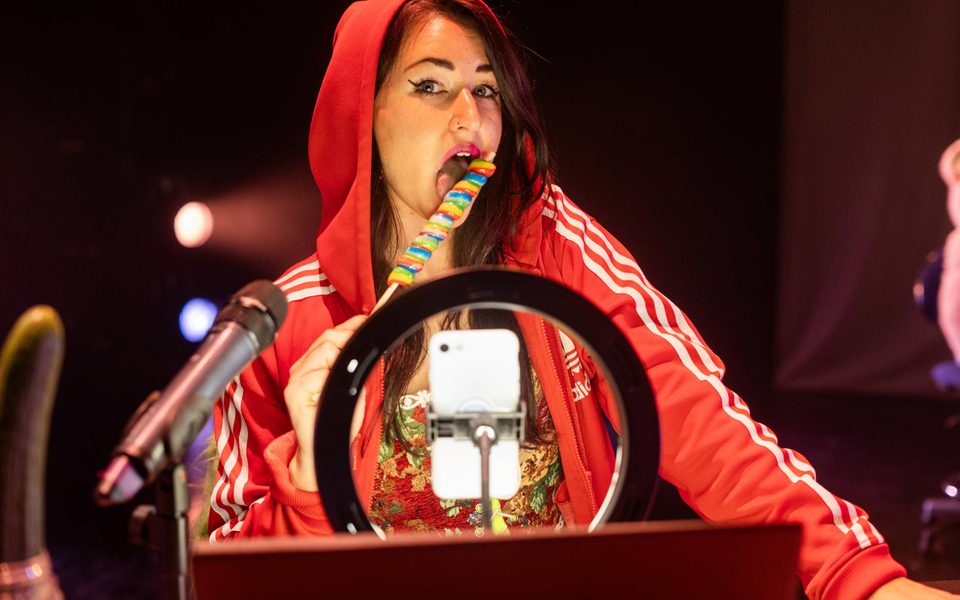 Louise Orwin, a woman with black hair and green eyes, is sitting behind a ring light and phone on a tripod with a microphone on her right. She is wearing a bright pink tracksuit and is licking a rainbow lollipop while looking into the camera.