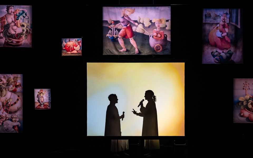 In the centre, two people stand behind a pastel-coloured screen, their black silhouettes projected onto it, they are facing each other and gesticulating each with a microphone in hand. A black wall behind features abstract paintings featuring nude doll-like female characters painted in reds, tans and greens.