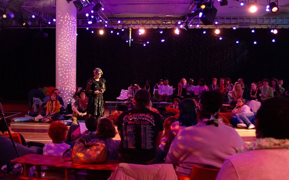 Artist Stacy Makishi, a woman with a short grey bob and glasses who is wearing a black frilled shirt with mesh sleeves and a black skirt with tights, stands in the middle of a relaxed yet tentative crowd. The crowd are all wearing Hawaiian-inspired flower wreaths around their neck. The room is warmly lit with pink and stippled colours from the visible lighting rigs on the celling.